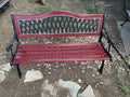 A red and black metal bench with a decorative lattice backrest, photographed outdoors on a dirt and gravel surface.